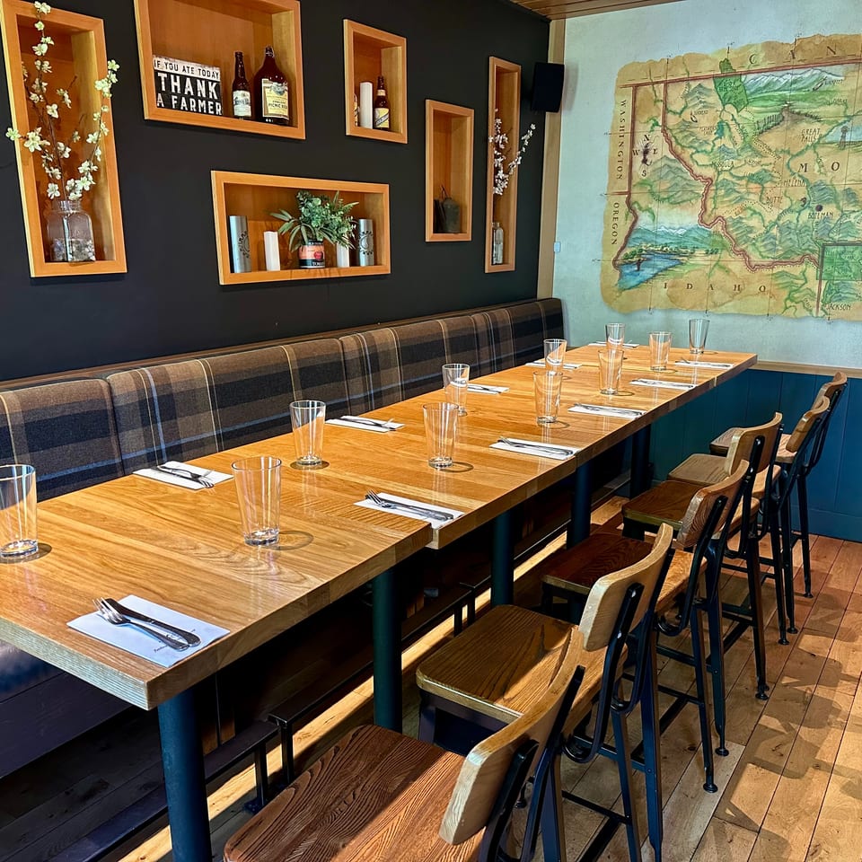Long wooden restaurant table set with glasses and cutlery, with a plaid banquette and map on the wall.