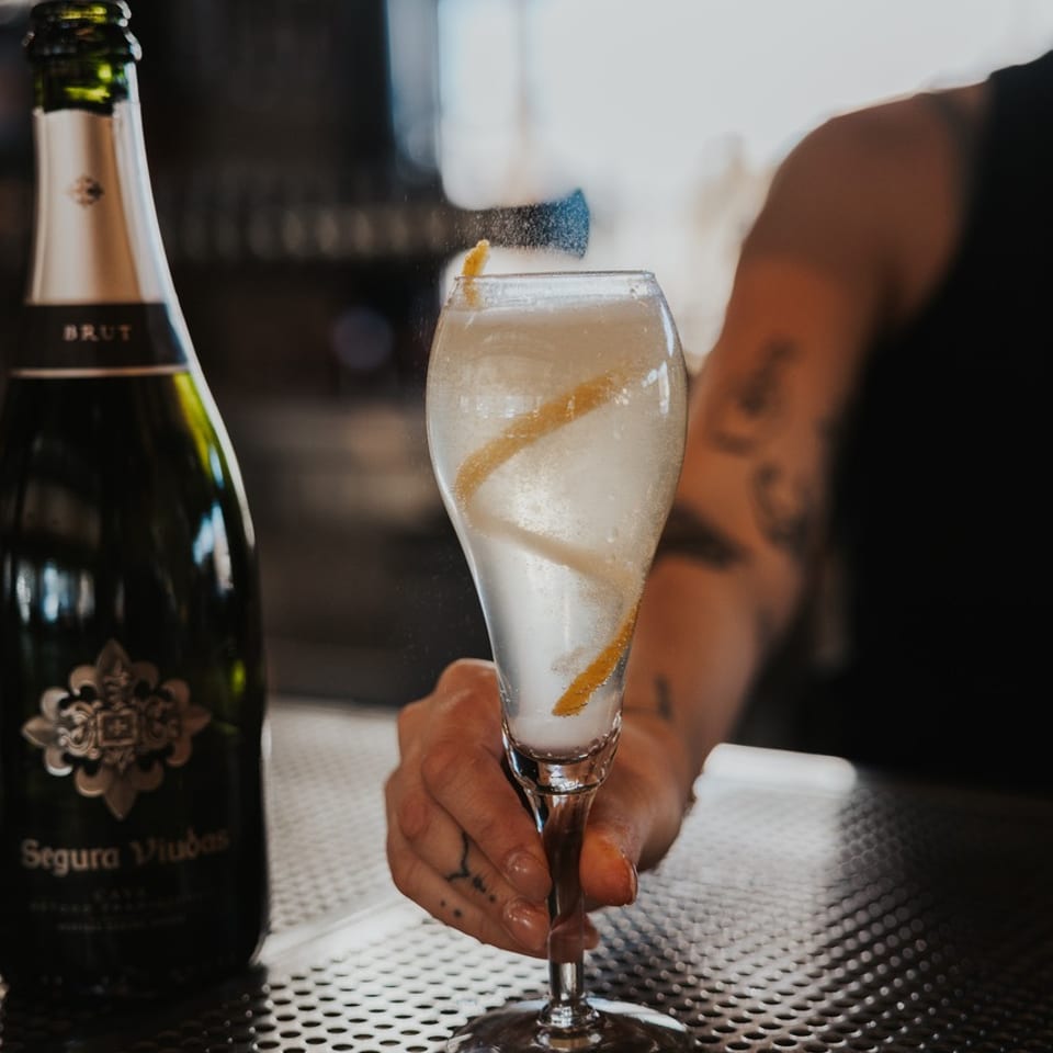 A hand holding a sparkling cocktail with a lemon twist next to a bottle of Segura Viudas Brut.