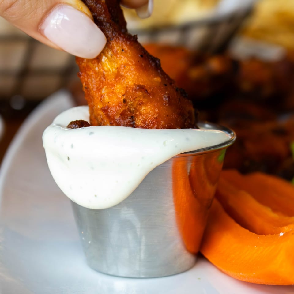 Hand dipping a crispy chicken wing into a metal cup of creamy white sauce, with carrot sticks.