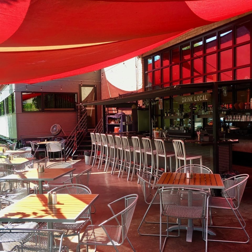 Restaurant patio with red shade sails, metal tables, a bar, and a train car building.