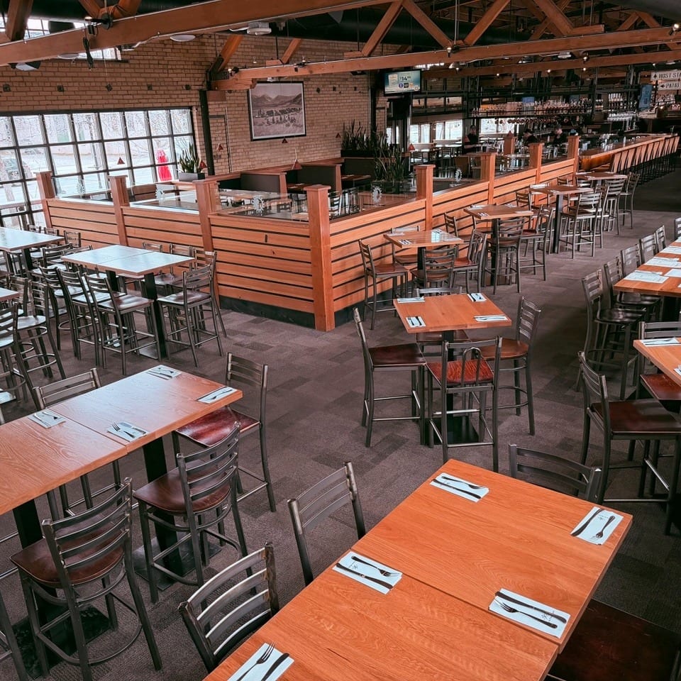 Empty restaurant dining room with numerous tables, high chairs, a bar, and large windows.