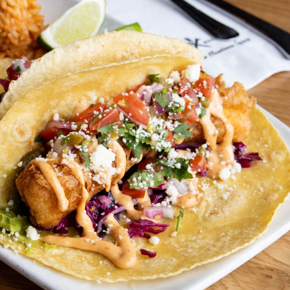 A close-up of a fish taco with red cabbage, pico de gallo, and a creamy sauce.