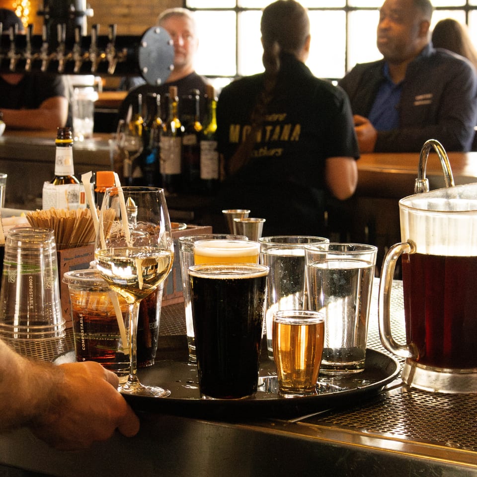 Bartender's hand holds a tray of beer, wine, and water glasses at a bustling bar.