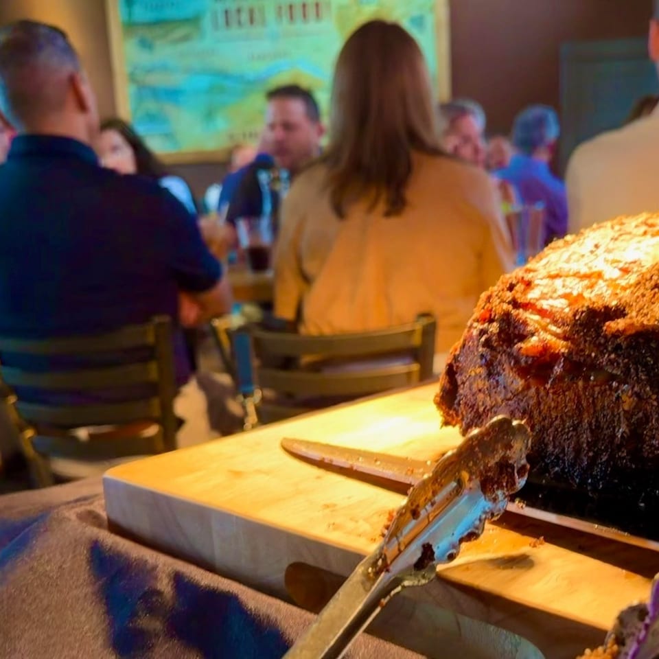 A large roasted meat sits on a carving board with tongs, in front of blurred restaurant patrons.