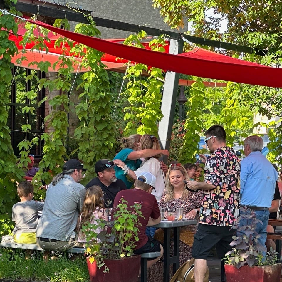 Patrons enjoying an outdoor patio with red awnings and lush green vines, two women embracing.