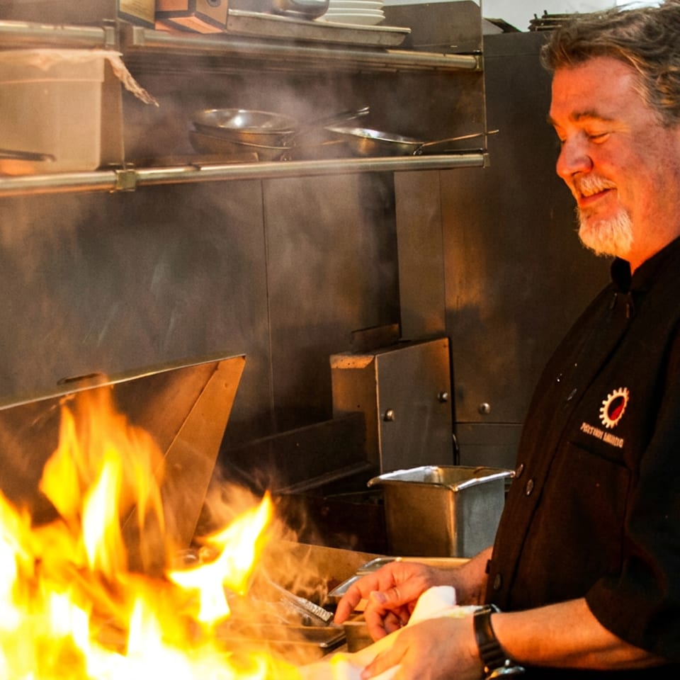 Chef in a black uniform smiling while cooking over a flaming grill in a commercial kitchen.