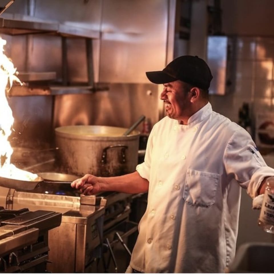 Smiling chef flambéing food with large flames, holding a Lainey's Vodka bottle in a restaurant kitchen.