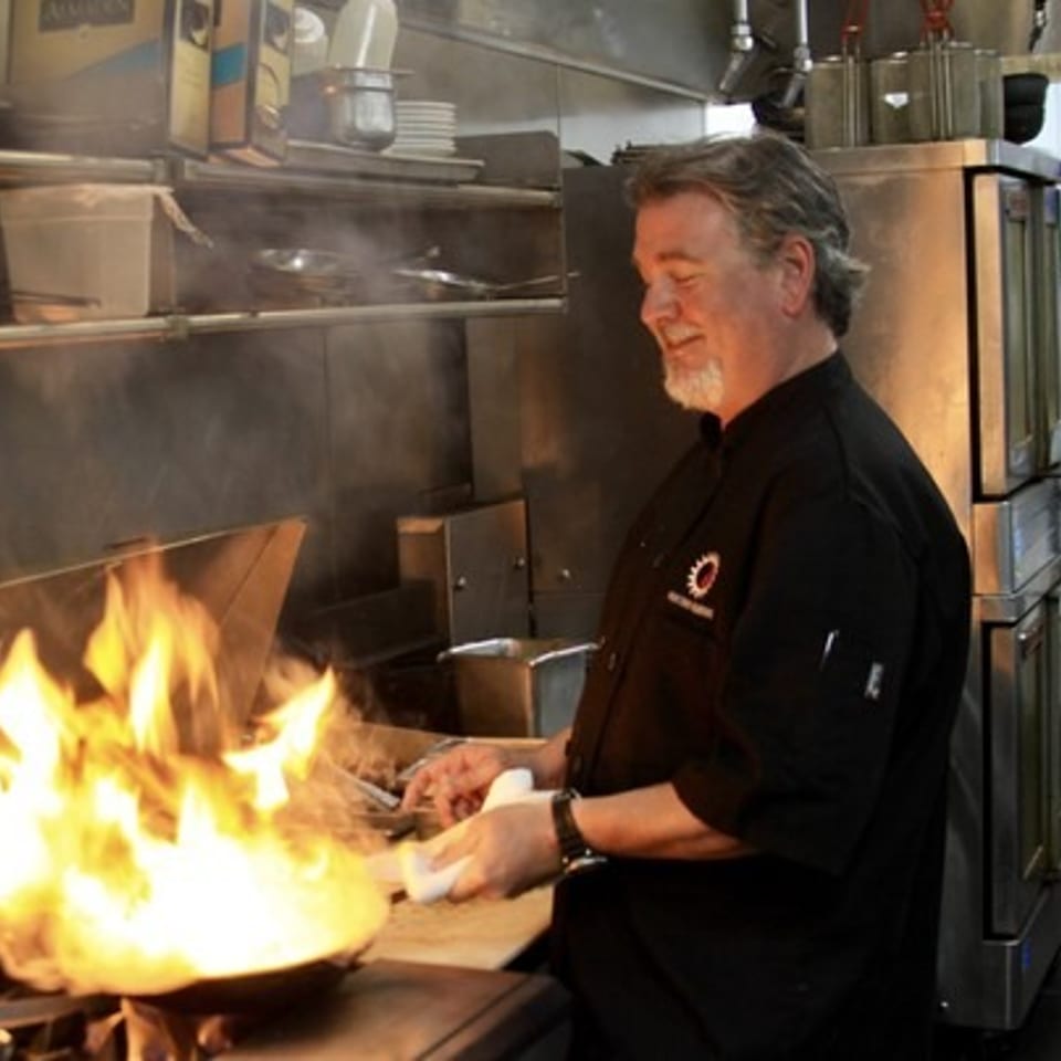 Smiling chef in a black uniform cooks with large flames rising from a pan in a commercial kitchen.