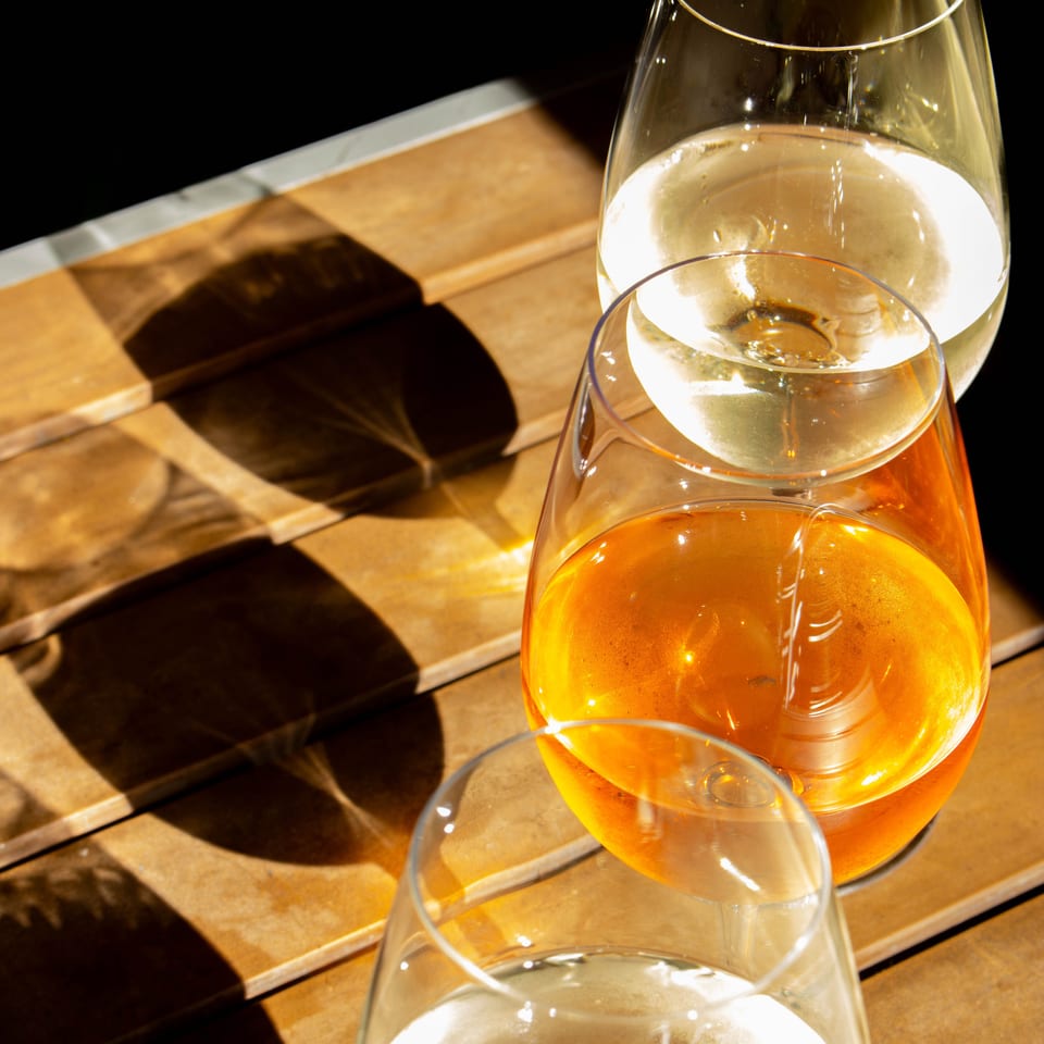 Three wine glasses, two white and one orange, sit on a sun-drenched wooden table.
