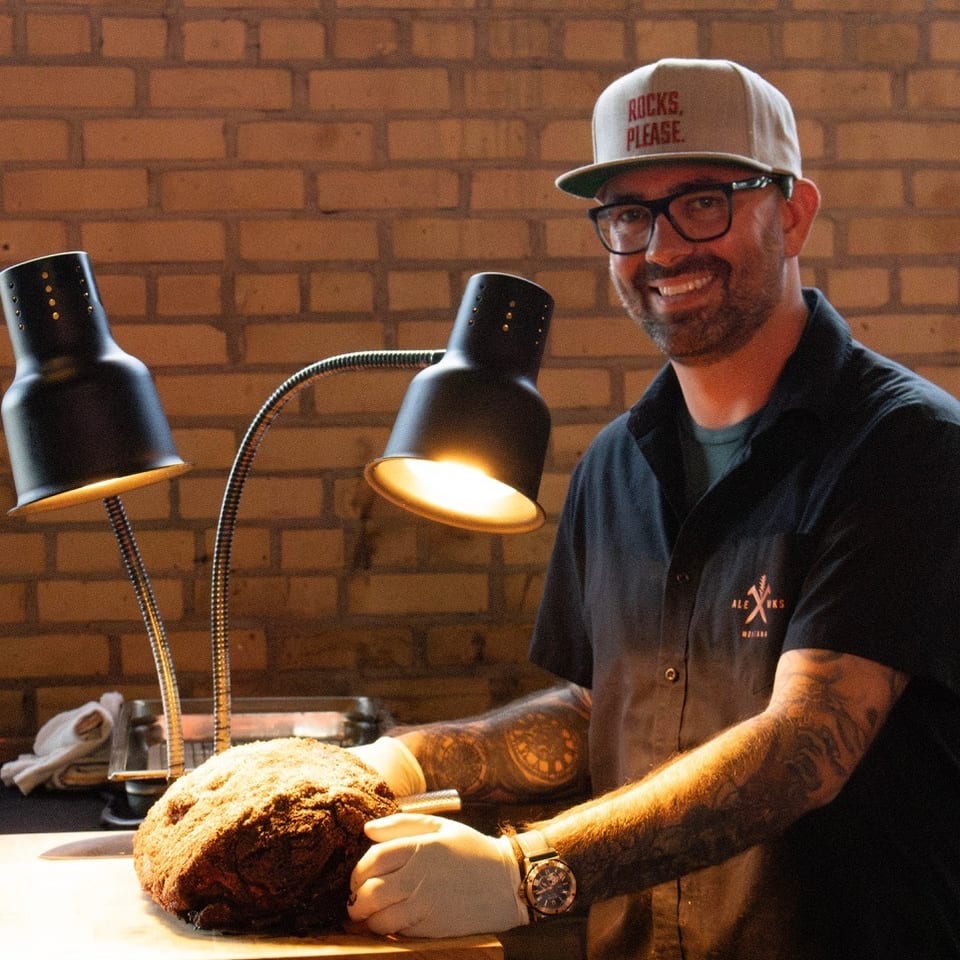 Chef in a hat and glasses smiles while carving a large roast under heat lamps.