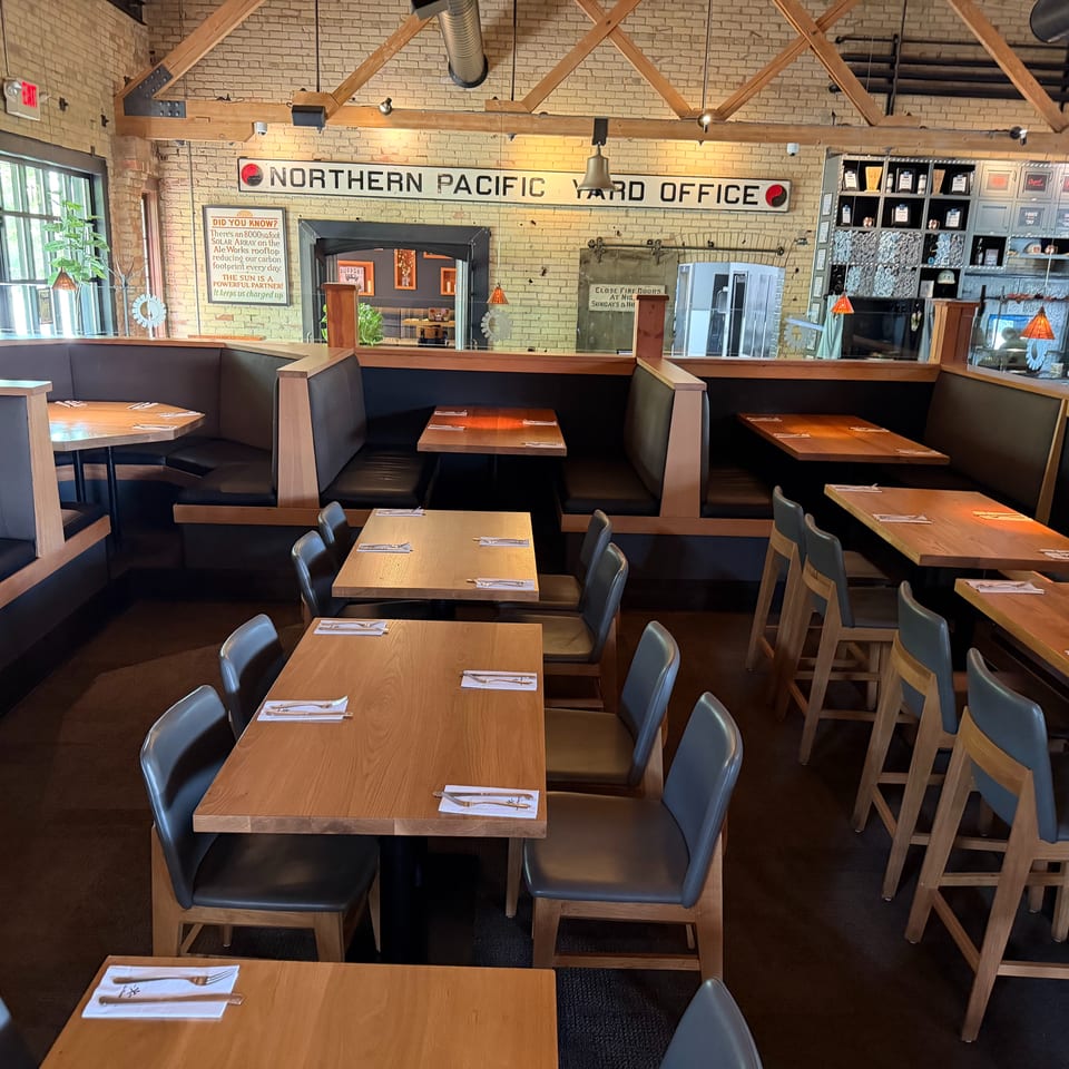 Industrial-style restaurant interior with wooden tables, dark booths, brick walls, and a "Northern Pacific Yard Office" sign.