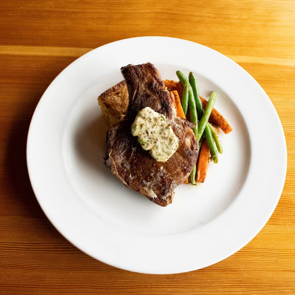 Grilled steak with herb butter, green beans, carrots, and Yorkshire pudding on a white plate.