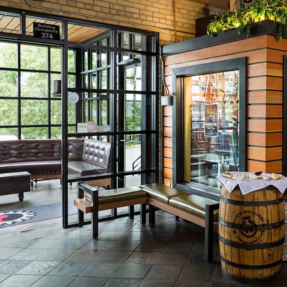 Modern restaurant entrance with glass walls, leather seating, and a sampling station next to a wooden barrel.