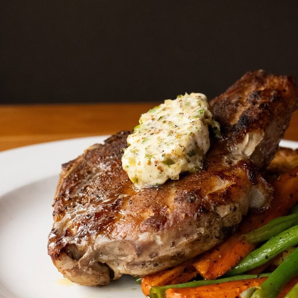 Grilled steak topped with compound butter, served with roasted carrots and green beans on a white plate.
