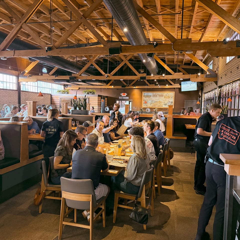 Diners enjoy meals in a bustling restaurant with exposed wooden beams and large windows.