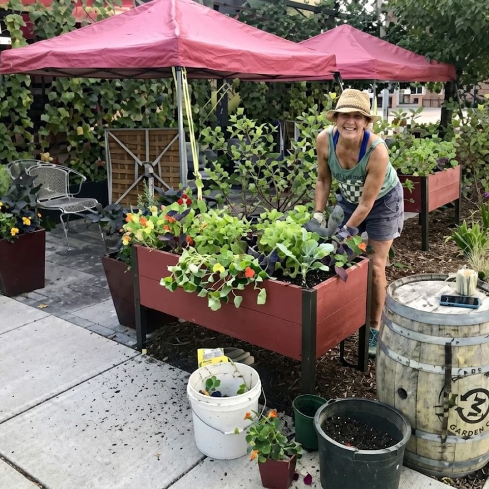 Smiling woman in a hat gardening in raised beds under red awnings at a restaurant.