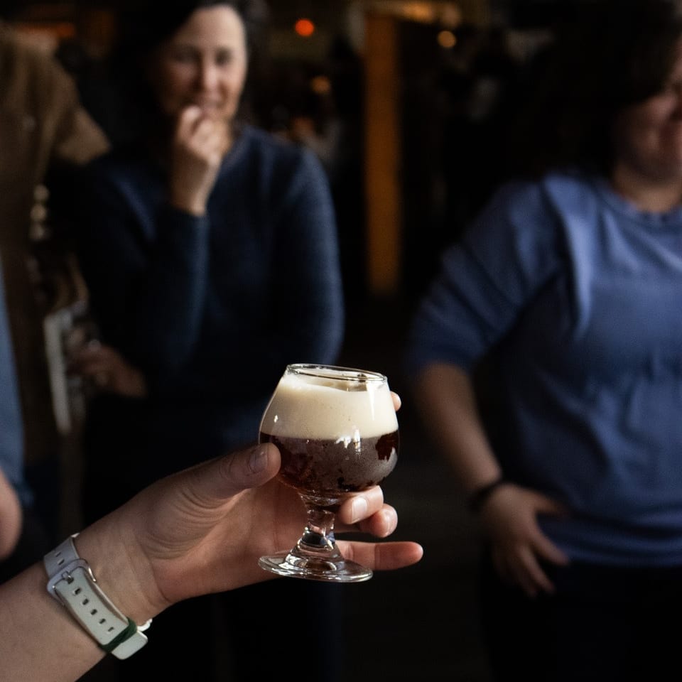 A hand holds a small glass of dark beer with a foamy head, with blurred people in the background.