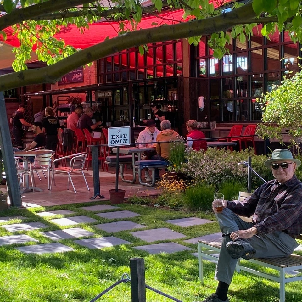 Older man enjoying a drink on a bench in a garden area next to a lively restaurant patio.