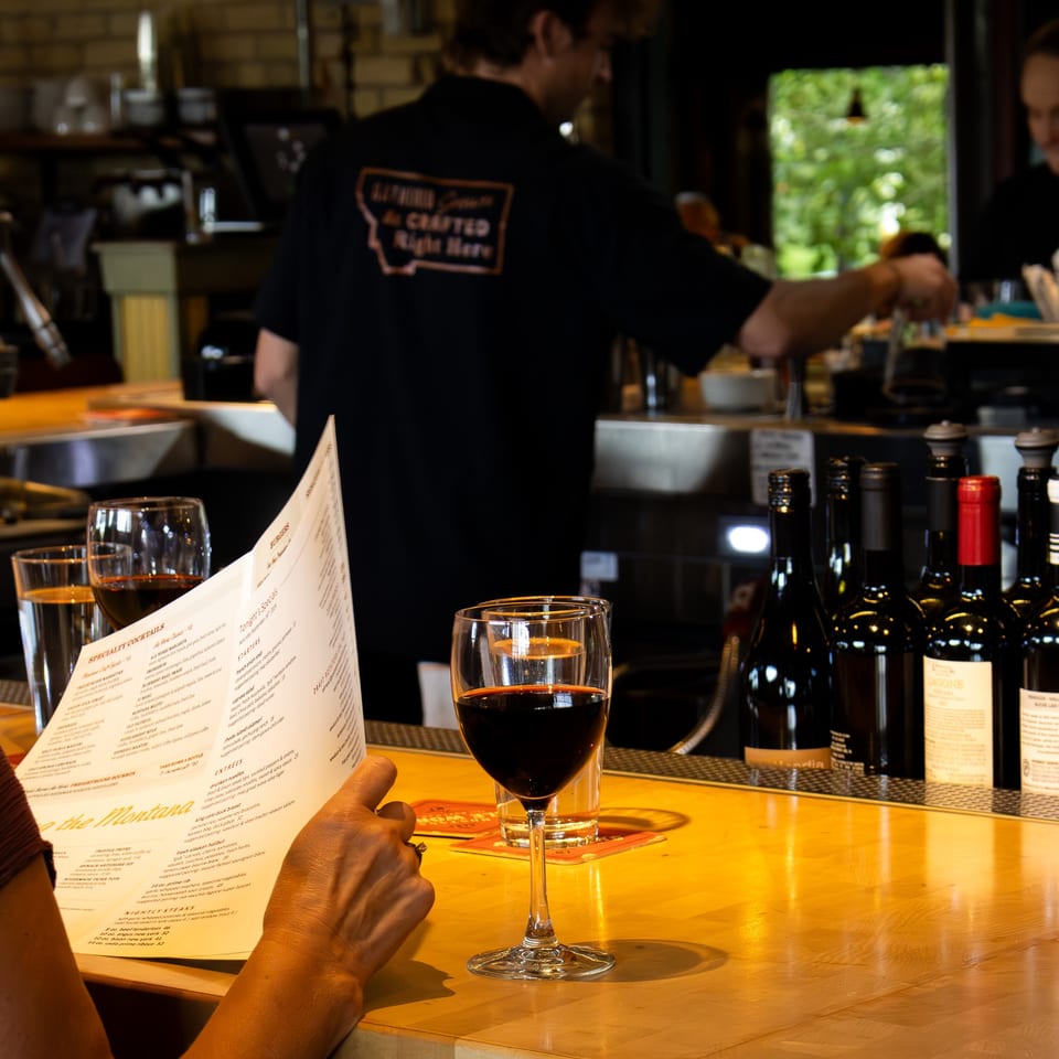 A customer reads a menu at a bar with a glass of red wine and a bartender.