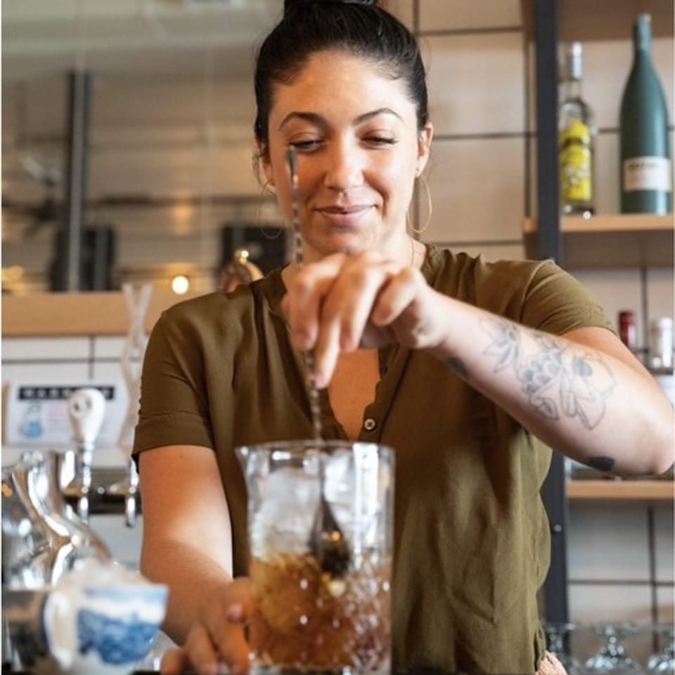 Smiling bartender stirring a drink with ice in a mixing glass behind a bar.