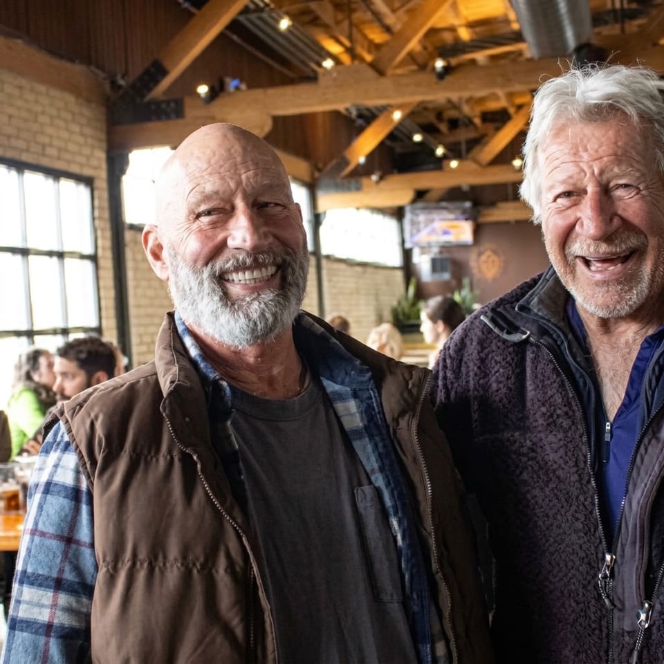 Two smiling older men, one with a beard, standing in a lively restaurant with exposed beams.