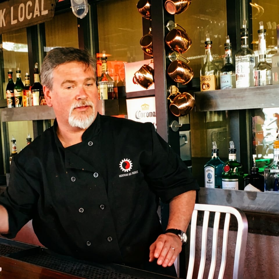 A chef in a black coat stands behind a bar with liquor bottles and a "DRINK LOCAL" sign.