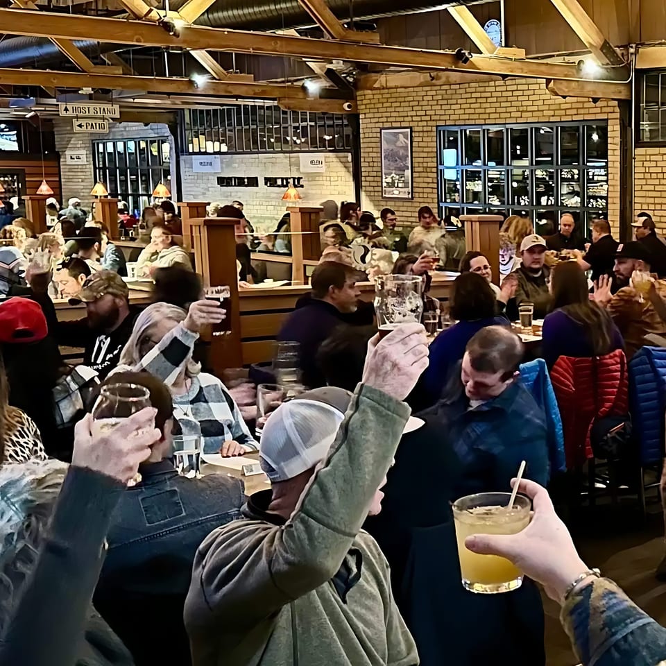 Diners raise their glasses in a toast inside a bustling restaurant with brick walls and wooden beams.
