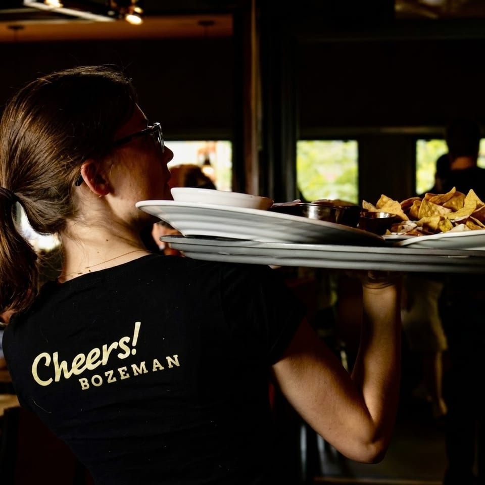 Waitress in a "Cheers! Bozeman" shirt carrying a tray of nachos and dips.