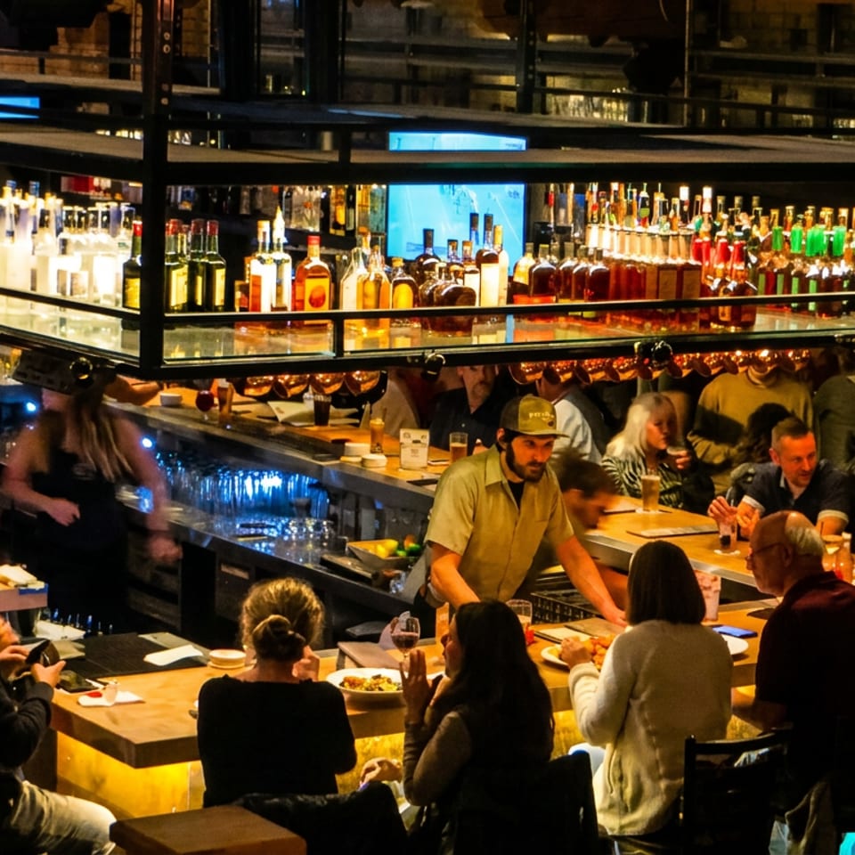 A lively restaurant bar scene with patrons seated at the counter and tables, and bartenders working.