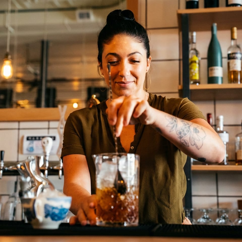 A smiling bartender with a tattooed arm stirs a cocktail in a mixing glass at a bar.