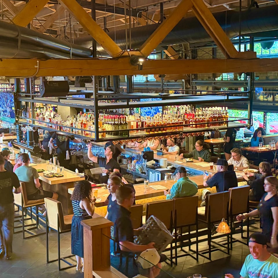 Lively restaurant interior with a long bar, many customers, and high ceilings with exposed beams.